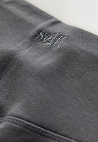 Gråt stof med en glat tekstur, med en broderet logo med teksten ".self." i en lidt mørkere nuance, placeret på linningen.