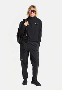 Completo sportivo nero con maglia a maniche lunghe con cerniera a un quarto e pantaloni a taglio largo, entrambi con dettagli logo, abbinati a grandi occhiali da sole alla moda.