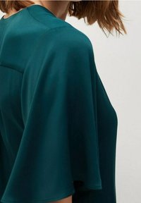 Spalla e parte superiore del torso di una donna che indossa una blusa verde scuro con maniche corte ampie e fluenti, e capelli castano chiaro.