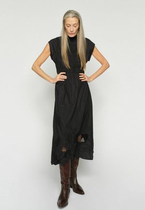 Žena s dlhými sivými vlasmi, oblečená v čiernych midi šatách bez rukávov s kvetinovými výšivkami a hnedými čižmami po kolená, stojaca s rukami v bok.