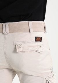 Beige cargobroeken van een gestructureerde stof, met een brede beige tailleband en een opgestikte zak aan de achterkant met een label eraan bevestigd.