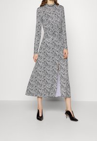 Eine Frau trägt ein schwarz-weiß gestreiftes Langarm-Midi-Kleid mit einem Schlitz vorne und schwarzen spitzen High Heels, während sie vor einem schlichten Hintergrund steht.