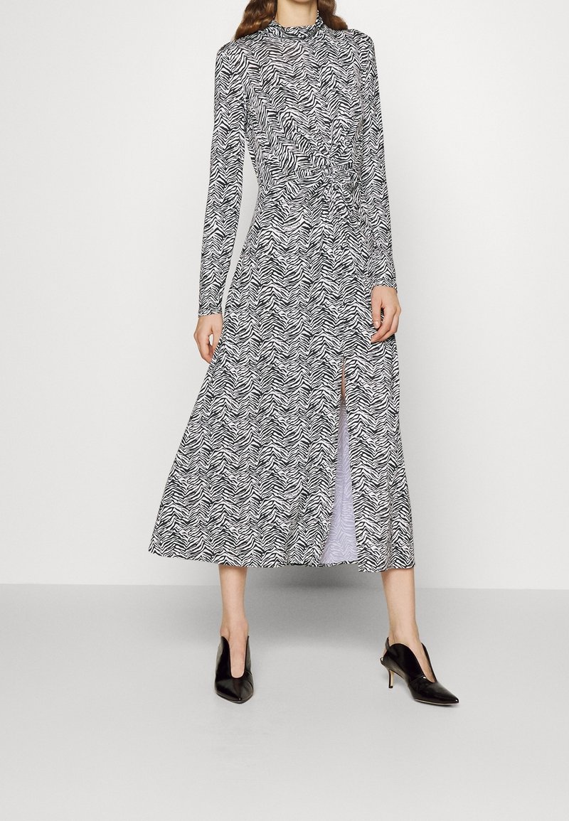 Eine Frau trägt ein schwarz-weiß gestreiftes Langarm-Midi-Kleid mit einem Schlitz vorne und schwarzen spitzen High Heels, während sie vor einem schlichten Hintergrund steht.