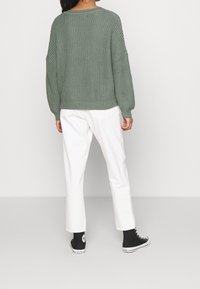 Maglione verde a maglia con spalle cadenti, texture a coste e vestibilità ampia, abbinato a pantaloni bianchi a gamba dritta e sneakers nere.