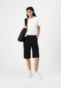 Chemise blanche à manches courtes avec un design en œillets, associée à un short noir jusqu'aux genoux. Le modèle porte un sac rembourré noir et des baskets blanches.