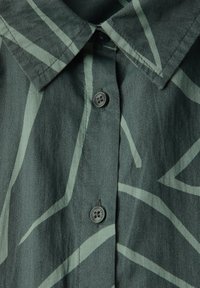 Camicia button-up in tessuto verde scuro con motivi a linee astratte verde chiaro; presenta un colletto e due bottoni visibili.