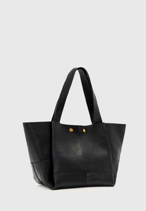 Czarna skórzana torba typu tote z szerokimi uchwytami i dwoma małymi złotymi guzikami z przodu, na jednolitym jasnoszarym tle.