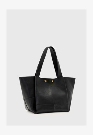 Czarna skórzana torba typu tote z szerokimi uchwytami i dwoma małymi złotymi guzikami z przodu, na jednolitym jasnoszarym tle.