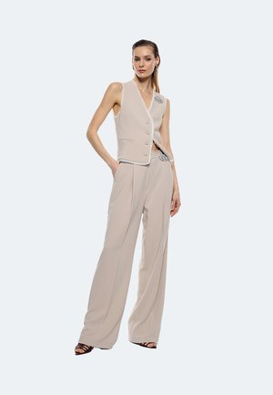 Gilet beige su misura con scollo a V, dettagli dei bottoni e accentuazione con spilla abbinato a pantaloni a gamba larga. Tessuto liscio con una vestibilità strutturata.