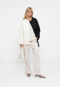 Mulher está de pé contra um fundo branco, vestindo uma jaqueta com blocos de cores em preto e branco, calças bege, saltos taupe e segurando uma pequena bolsa branca.
