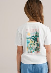 Camiseta blanca de algodón con un gráfico retro colorido de palmera en la parte posterior, con el texto "RIVIERA" y "CANNES 1982". Mangas cortas, corte relajado.