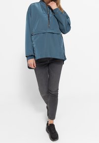Lightweight blue pullover jacket with a half-zip, elastic cuffs, and adjustable hem; worn with gray slim-fit jeans and black sneakers.