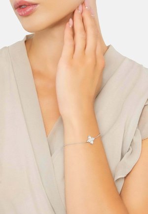 Silberarmband mit floraler Gestaltung, das drei klare Edelsteine enthält, am Handgelenk vor einem hellbeigen Stoffhintergrund getragen.