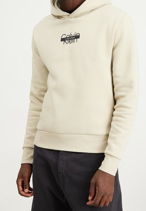 Sweat-shirt à capuche beige en tissu doux. Présente des poignets et un ourlet côtelés. Logo Calvin Klein noir imprimé sur la poitrine.