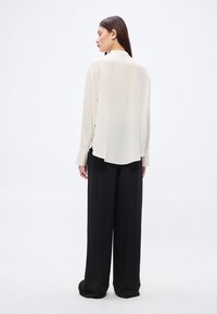 Világos bézs hosszú ujjú blúz ívelt szegéllyel és mandzsettás ujjakkal, fekete, széles szárú nadrággal párosítva. Simaságot sugárzó textúra, minimalista dizájn.