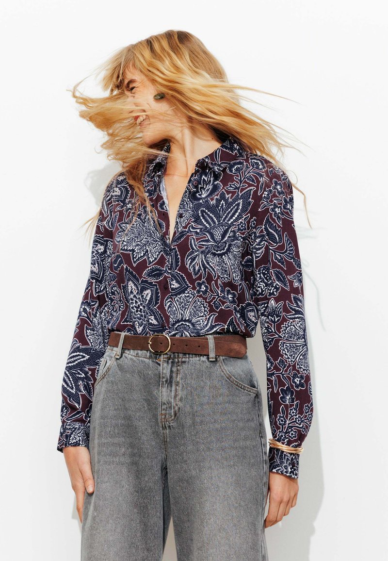 Chemise à manches longues avec un motif floral bleu marine sur fond bordeaux, associée à un pantalon en jean taille haute gris clair et une ceinture marron.