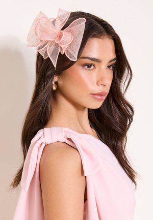 Žena s dlhými tmavými vlasmi, ktorá má na sebe svetloružové šaty s mašľou na ramene a veľkým jemným ružovým sieťkovým doplnkom do vlasov na strane.