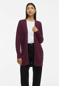 Burgunderfarbener Strickcardigan mit strukturiertem Design, offener Front, langen Ärmeln und zwei Fronttaschen; getragen über einem weißen Hemd und schwarzen Hosen.