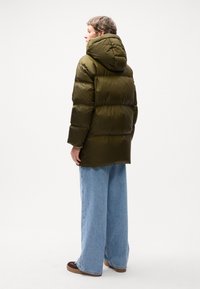 Jachetă puffer de culoare verde măslinie cu glugă, cu quilturi orizontale. Asortată cu blugi evazați de un albastru deschis și încălțăminte maro.