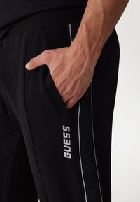 Schwarze Sporthosen mit schlanker Passform, ausgestattet mit einem weißen "GUESS"-Logo, Seitentasche und strukturiertem schwarzen Seitestreifenmuster.