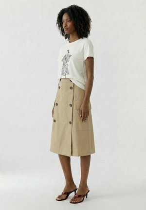 Femme aux cheveux bouclés portant un t-shirt blanc à motif, une jupe midi beige boutonnée et des sandales à talons marron, debout de profil.