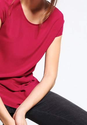 Osoba ubrana w czerwony krótki rękaw i ciemne dżinsy, siedząca z rękami spoczywającymi na kolanach, na białym tle.