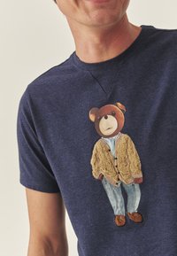 Tumši zils T-krekls ar izšūtu lāci, kas valkā bēša krāsas kardiganu, gaišu kreklu, zilas džinsas un brūnas kurpes. Mīksts auduma tekstūra.