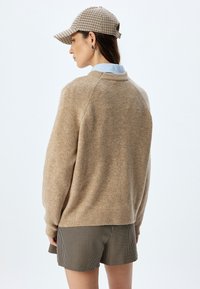 Beige Wollpullover mit Rundhalsausschnitt, kombiniert mit karierten Shorts. Die Mütze ist kariert mit einem gebogenen Schirm und einem verstellbaren Hinterband.