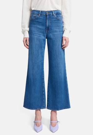 Weit geschnittene, hoch taillierte blaue Jeans aus Denim mit glatter Textur, getragen von einer Person in hellfarbigen Schuhen.