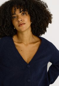 Cárdigan azul marino hecho de tela de punto suave, con escote en v profundo, dos botones redondos y mangas largas con un corte relajado.