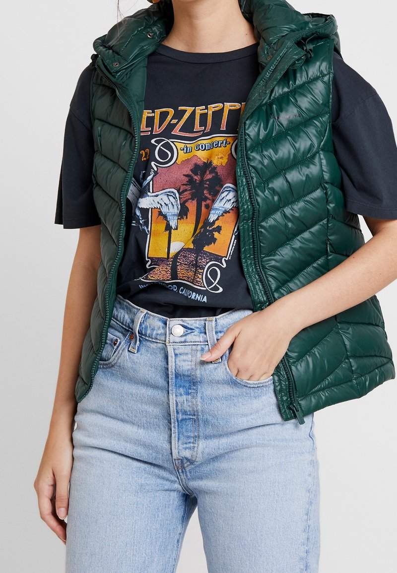 Osoba ubrana w zieloną kurtkę typu puchowego na czarnym t-shircie z grafiką Led Zeppelin i jasnoniebieskich jeansach z wysokim stanem, z jedną ręką w kieszeni.