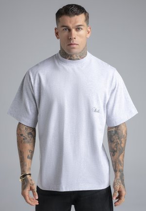 Homme avec tatouages au cou et au bras portant un t-shirt ample gris clair uni, debout devant un fond gris neutre.