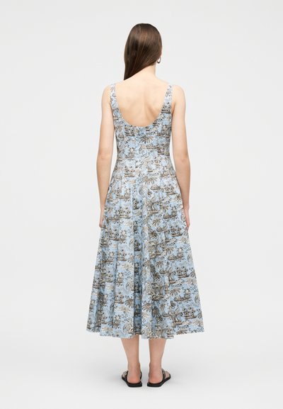 Γυναίκα που στέκεται με την πλάτη στραμμένη στην κάμερα, φορώντας ένα αμάνικο, ανοιχτό γαλάζιο φόρεμα με καφέ λουλουδάτο και φυσικό σχέδιο, και μαύρα σανδάλια.