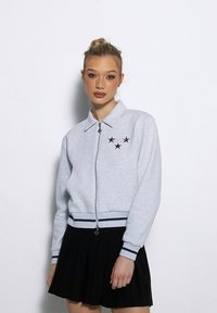 Szary zamek-suwakowy sweatshirt z kołnierzykiem, ozdobiony haftem czarnych gwiazd i prążkowanym wykończeniem na mankietach i dole, w zestawieniu z czarną spódnicą plisowaną.