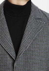 Grau und marineblauer Blazer im Hahnentritt-Muster, mit Schalkragen und strukturiertem Stoff, über einem schwarzen Rollkragenpullover getragen.