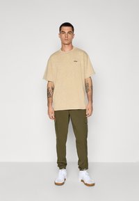 Beige crew-neck t-shirt with a subtle texture and small logo, paired with olive green cargo pants and white sneakers with brown soles.