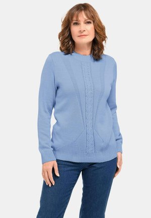 Donna con capelli castani fino alle spalle, indossa un maglione di lana leggero di colore azzurro con un motivo sulla parte anteriore e jeans di colore blu scuro, in piedi contro uno sfondo bianco.