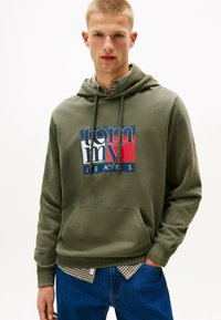 Oliven grøn hættetrøje med en forlommel, prydet med et blåt og rødt grafisk design, der står "tommy JEANS." Båret over en ternet skjorte.
