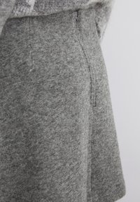 Close-up van een grijs wollen jas met textuur, die de zijrits en zakdetails toont tegen een neutrale achtergrond.