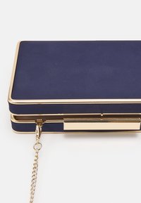 Navy blue clutch bag with a gold metal frame, featuring a textured surface, chain strap, and clasp closure. Compact rectangular shape.