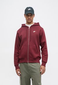 Fleece com capuz em tom bordeaux, com fecho, apresentando um logo branco da Nike no peito, combinado com calças cargo olivas e um chapéu preto.
