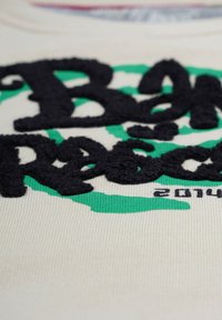 Puuvillainen t-paita, jossa on tekstuurinen musta ja vihreä graafinen kuvio, jossa lukee "Ban Rascal" ja alareunassa vuosi "2014", vaalean kermanvärisellä taustalla.