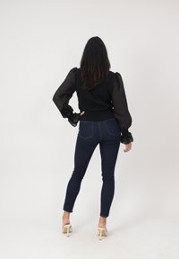Mujer de pie de espaldas vistiendo una blusa negra transparente de manga larga, jeans ajustados oscuros y sandalias blancas de tacón alto sobre un fondo claro y liso.
