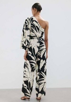 Mujer con el pelo recogido en un moño, que lleva un mono de un solo hombro con estampado tropical en blanco y negro y sandalias marrones con tacón, de pie mirando hacia atrás.