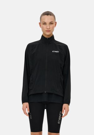 Woman wearing a black zip-up jacket and black knee-length cycling shorts with “CEP” logos, standing against a plain white background.