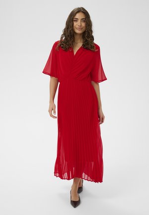 Kobieta o kręconych włosach ubrana w zwiewną, czerwoną, plisowaną sukienkę midi z krótkimi rękawami i czarne, szpiczaste szpilki, stojąca na białym tle.