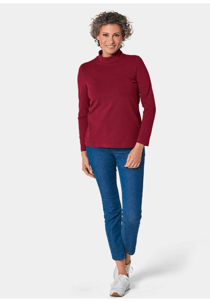 Bordeauxrood langemouwen top met hoge hals, gecombineerd met blauwe tapered jeans en witte sneakers. De stof lijkt zacht met een gladde textuur.