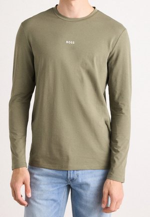Homme portant une chemise manches longues vert olive avec le texte "BOSS" centré sur la poitrine et un jean bleu clair, debout devant un fond uni.