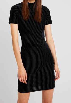 Frau trägt ein kurzes schwarzes Samtkleid mit vertikalen Streifen und kurzen Ärmeln, stehend vor einem schlichten hellen Hintergrund.