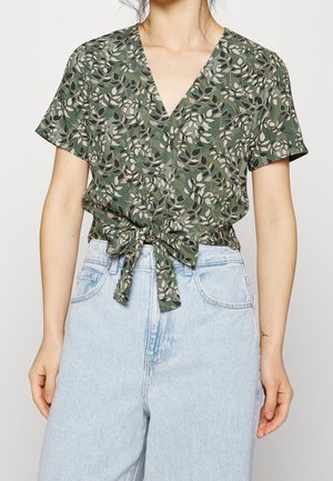 Blusa verde a fiori con scollo a V, maniche corte e un fiocco in vita, abbinata a jeans azzurri a vita alta.
